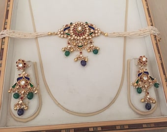 Op de afbeelding: Een gouden kettingset met een hanger en oorbellen. De hanger en oorbellen hebben een pauwenmotief met blauw, groen en rood email. De ketting is geregen met witte parels.