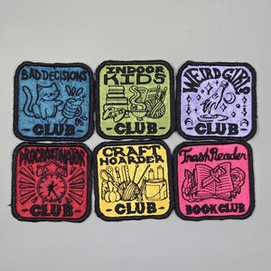 May include: Six embroidered patches with black borders. Each patch features a different design and the word "CLUB". Designs include a cat, books, a rocket, an alarm clock, craft supplies, and an open book.