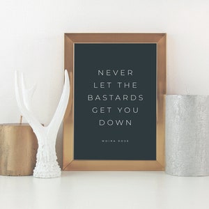 May include: A gold framed print with a black background and white text that reads "Never let the bastards get you down" with the name Moira Rose at the bottom. The print is sitting on a white surface with two candles and a white antler figurine.