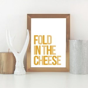 May include: A gold framed print with the text "FOLD IN THE CHEESE" in a yellow and orange colour scheme. The print is displayed on a white surface with two candles and a white antler decor.