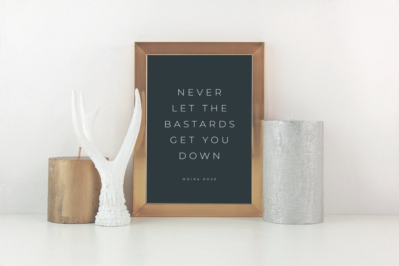 May include: A gold framed print with a black background and white text that reads "Never let the bastards get you down" with the name Moira Rose at the bottom. The print is sitting on a white surface with two candles and a white antler figurine.