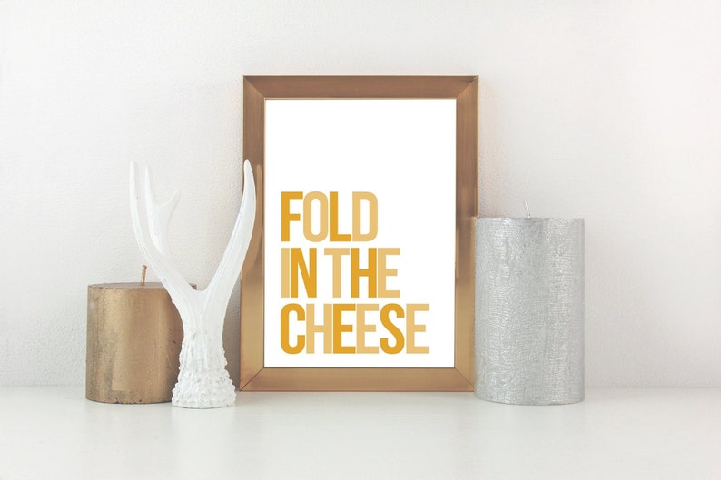 May include: A gold framed print with the text "FOLD IN THE CHEESE" in a yellow and orange colour scheme. The print is displayed on a white surface with two candles and a white antler decor.