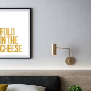 May include: A framed print with the text "FOLD IN THE CHEESE" in a gradient of yellow and orange. The print is hanging on a white wall above a bed with a black headboard.