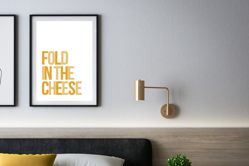 May include: A framed print with the text "FOLD IN THE CHEESE" in a gradient of yellow and orange. The print is hanging on a white wall above a bed with a black headboard.