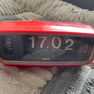 May include: A bright red rectangular flip clock with a black display panel showing the time 17.02 in white numerals. The word Rhythm is printed in white script below the time. A vertical numerical scale from 12 to 15 is visible on the left side.