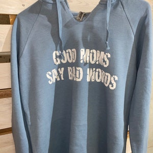 Op de afbeelding: Een lichtblauw hoodie met de tekst "Good Moms Say Bad Words" in wit gedrukt.