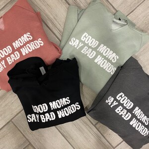 Op de afbeelding: Vier sweatshirts in verschillende kleuren met dezelfde tekst erop gedrukt: "Good Moms Say Bad Words". De sweatshirts zijn gevouwen en op een houten oppervlak gelegd.