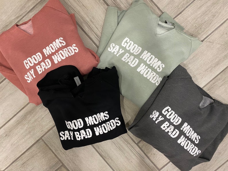 Op de afbeelding: Vier sweatshirts in verschillende kleuren met dezelfde tekst erop gedrukt: "Good Moms Say Bad Words". De sweatshirts zijn gevouwen en op een houten oppervlak gelegd.