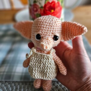 May include: A pink crocheted amigurumi toy shaped like a house elf with large ears, brown eyes, and a beige crocheted dress.