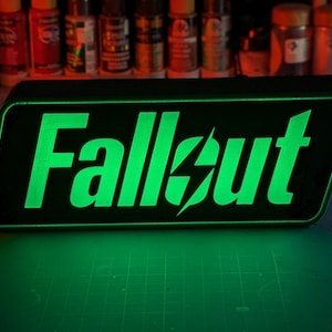 Könnte beinhalten: Ein schwarzes und grünes Neon-Schild mit dem Wort "Fallout" in einer stilisierten Schriftart mit einem Blitz.