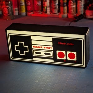 May include: A black and white Nintendo controller shaped light with red buttons. The light is on and illuminating the controller buttons.