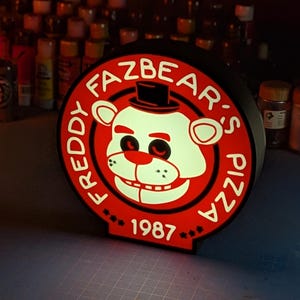 Może przedstawiać: Czerwono-biały znak 3D z napisem "Freddy Fazbear's Pizza" i rokiem "1987" w kółku. Znak przedstawia białego niedźwiedzia w czarnym cylindrze i z czerwonymi oczami.