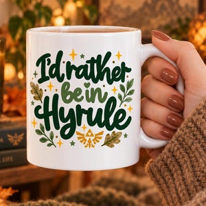 May include: A white ceramic mug featuring the dark green text "I'd rather be in Hyrule" in a cursive font. The design includes small green leaves, brown oak leaves, yellow star accents, and a gold-coloured Triforce symbol centred below the text.