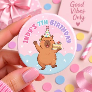 May include: A round button featuring a cartoon capybara wearing a colourful polka-dot party hat, holding a slice of cake. The background is light pink with blue clouds and white stars. The text reads INDY'S 7TH BIRTHDAY in purple and pink.