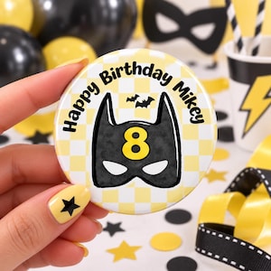 May include: A round button featuring a yellow and white checkered background with the text Happy Birthday Mikey in black. The centre displays a black superhero mask graphic with a yellow number 8 and two small black bats above the mask.