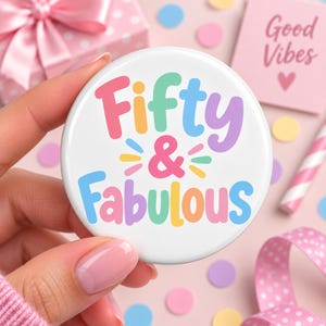 May include: A round white button featuring the colourful text Fifty & Fabulous in a playful font. The words are written in shades of pink, orange, green, blue, and purple, with small decorative dashes surrounding the ampersand symbol.