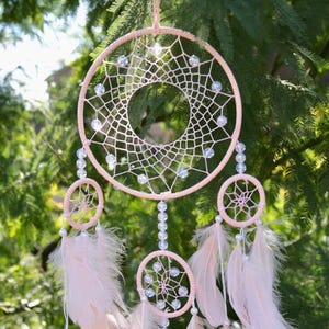 May include: A pink dream catcher with white beads and feathers. The dream catcher has a large circle in the center and three smaller circles below it.