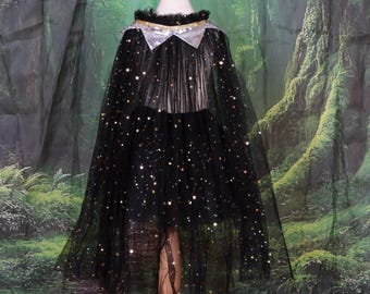 Puede incluir: Una capa de tul negro con detalles de estrellas doradas. La capa presenta un cuello plateado con ribete dorado y se muestra en un maniquí. El fondo es una escena de bosque.