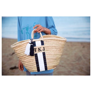 Peut inclure: Un grand sac cabas en paille tressée avec des rayures noires et blanches et un pompon noir. Le sac porte les initiales "TKJ" imprimées sur le devant.