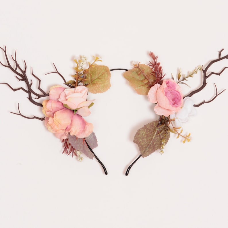 Puede incluir: Dos diademas decorativas con forma de cuernos de ciervo. Cada diadema presenta cuernos marrones, rosas rosas y melocot&oacute;n, y hojas verdes y marrones. Las diademas est&aacute;n sobre un fondo blanco.