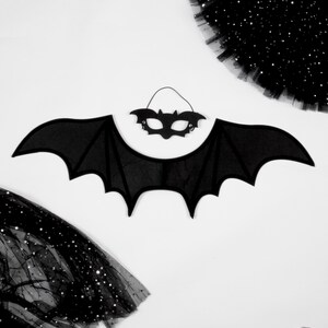 May include: A black bat wing costume accessory with a matching black bat-shaped eye mask. The wings feature a pointed, scalloped edge design, and the mask includes a thin elastic strap for securing it in place against the face.