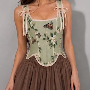 May include: A sage green corset top featuring a floral and monarch butterfly print with a square neckline and light pink ribbon shoulder ties. The bodice has a structured, paneled design with a scalloped hem, paired with a brown tulle skirt.