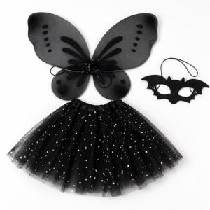 May include: A black costume set consisting of sheer fairy wings with glittery accents, a black bat-shaped eye mask with an elastic strap, and a black tulle tutu skirt featuring a pattern of small silver stars and crescent moons throughout.