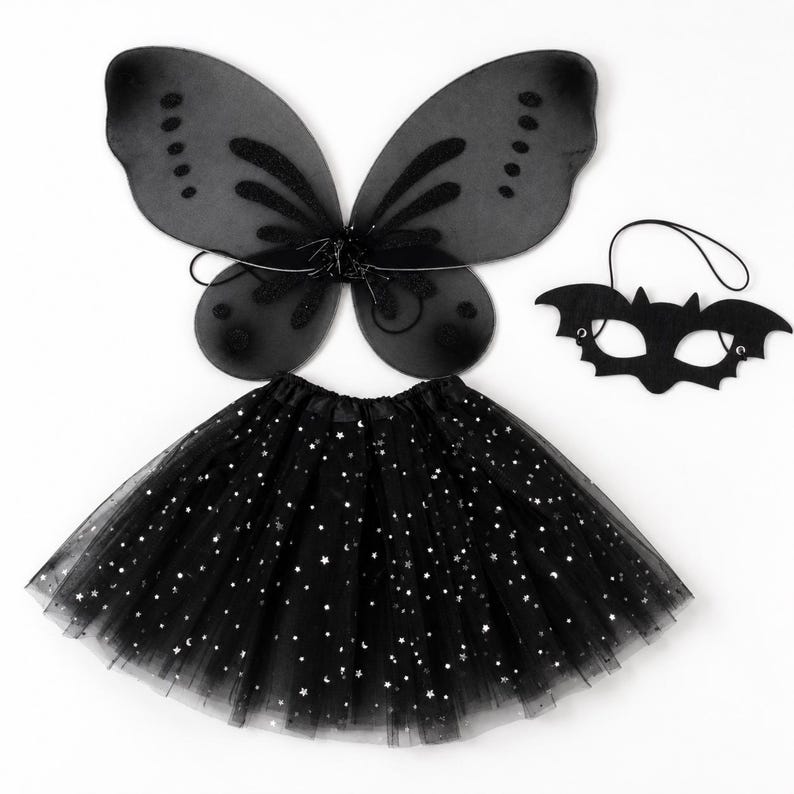 May include: A black costume set consisting of sheer fairy wings with glittery accents, a black bat-shaped eye mask with an elastic strap, and a black tulle tutu skirt featuring a pattern of small silver stars and crescent moons throughout.