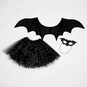 May include: A black bat costume set consisting of a pair of fabric bat wings, a black tulle skirt featuring silver star-shaped sequins, and a black felt bat-shaped eye mask with an attached elastic cord for securing the mask in place.