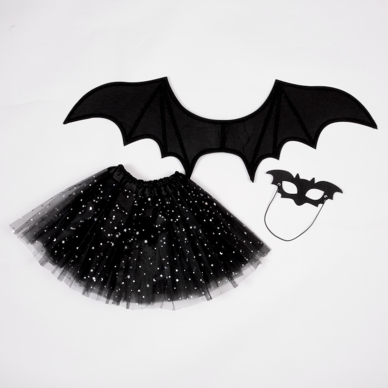 May include: A black bat costume set consisting of a pair of fabric bat wings, a black tulle skirt featuring silver star-shaped sequins, and a black felt bat-shaped eye mask with an attached elastic cord for securing the mask in place.
