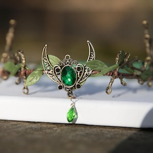 Könnte beinhalten: Eine grüne Edelsteinkrone mit einem Halbmond-Design, verziert mit künstlichen Blättern und einem herabhängenden grünen Edelstein-Tropfen.