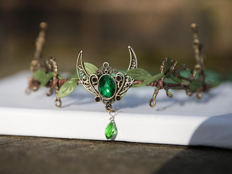 Op de afbeelding: Een groene edelsteenkroon met een halve maan ontwerp, versierd met nepblad en een hangende groene edelsteendruppel.