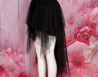 May include: A black tulle skirt with a high-low hemline. The skirt features multiple layers of sheer fabric, creating a voluminous effect. The waistband is elasticized. The skirt is displayed on a mannequin against a pink floral backdrop.