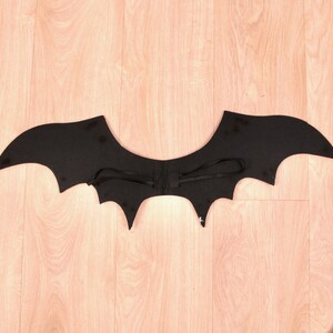 May include: A pair of black bat wings featuring a scalloped edge design. The wings are constructed from a solid black fabric and include two thin black elastic straps attached to the centre, designed to be worn over the shoulders for support.