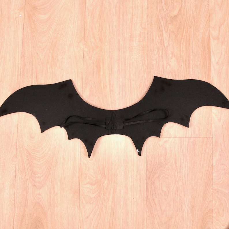 May include: A pair of black bat wings featuring a scalloped edge design. The wings are constructed from a solid black fabric and include two thin black elastic straps attached to the centre, designed to be worn over the shoulders for support.