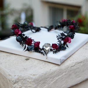May include: A gothic-style floral crown featuring black leaves and dark red roses. A silver-colored bird skull is centered on the crown. The crown is displayed on a white surface.