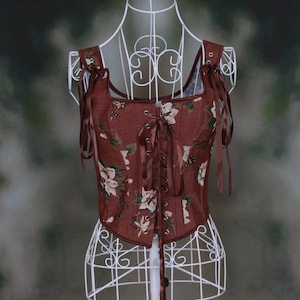 May include: A brown corset top featuring a floral pattern with white flowers and green leaves. The garment has a square neckline, a front lace-up closure with brown ribbon, and shoulder straps with adjustable brown ribbon ties.