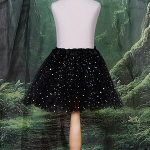 May include: A black tulle skirt featuring a gathered elastic waistband and a pattern of small, shimmering silver stars and crescent moons scattered across the layered fabric, creating a voluminous silhouette on a display mannequin.