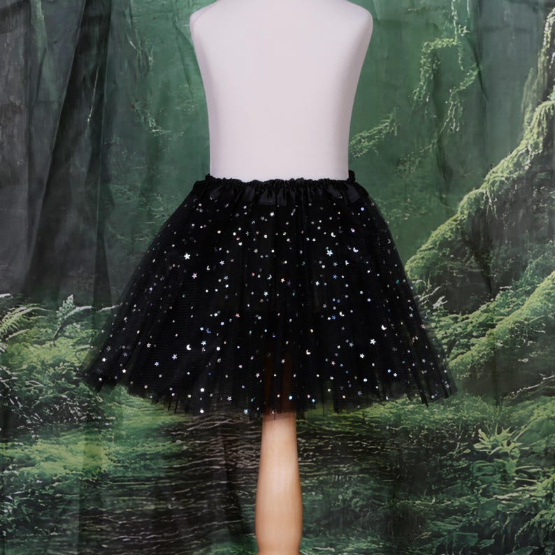 May include: A black tulle skirt featuring a gathered elastic waistband and a pattern of small, shimmering silver stars and crescent moons scattered across the layered fabric, creating a voluminous silhouette on a display mannequin.
