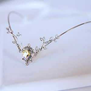 May include: A silver-toned metal headpiece featuring a central oval-shaped iridescent crystal set in an ornate frame, flanked by symmetrical branches with small leaf-like details extending outward along a thin, curved wire band.