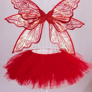 May include: A red fairy costume set, featuring a tulle tutu skirt and matching butterfly wings. The wings are red with glitter accents and a delicate pattern. The costume is displayed on a white mannequin.