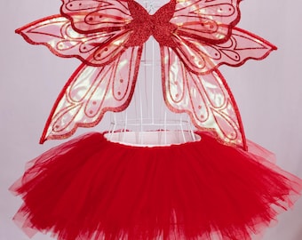 May include: A red fairy costume set, featuring a tulle tutu skirt and matching butterfly wings. The wings are red with glitter accents and a delicate pattern. The costume is displayed on a white mannequin.