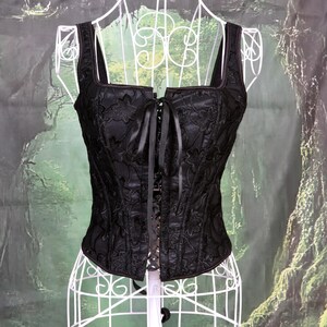 Peut inclure: Un corset en dentelle noire avec un décolleté carré et un laçage sur le devant. Le corset est sur un mannequin blanc et a un ruban de satin à nouer en haut.