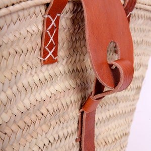 Può includere: Un cesto in paglia intrecciata con fibre naturali color beige e cinghie in pelle marrone. I manici in pelle sono fissati al corpo del cesto con cuciture a croce bianche e una linguetta di chiusura in pelle marrone pende dal bordo.