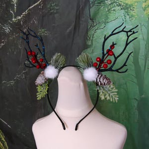 May include: A black headband featuring decorative antlers, red berries, white pom-poms, pine cones, and green foliage. The antlers are black with blue detailing. The headband is designed with a festive winter or holiday theme in mind.