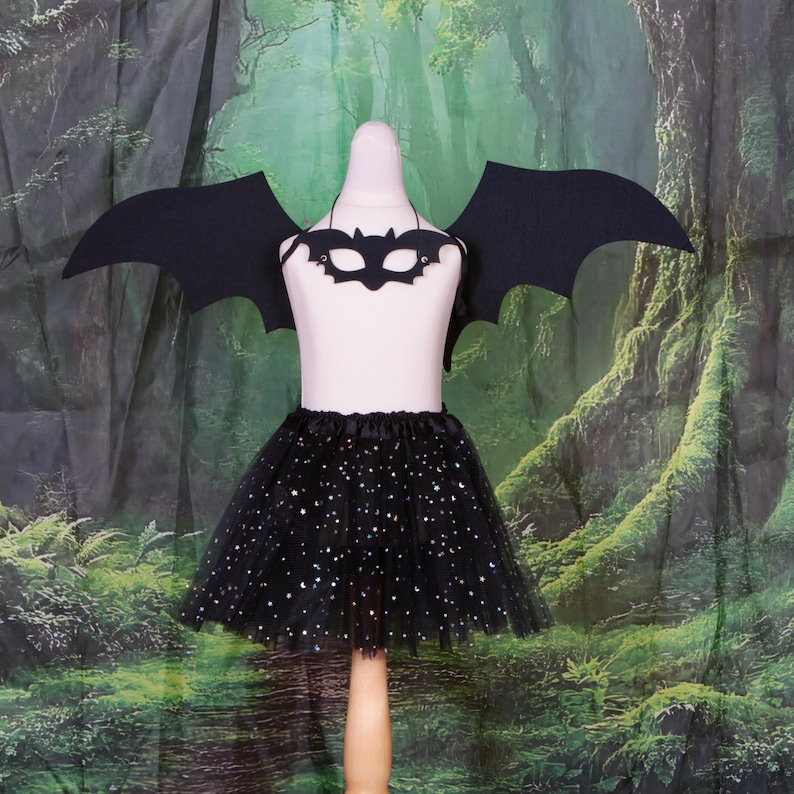 May include: A black bat costume set featuring a pair of large, pointed bat wings, a matching bat-shaped eye mask, and a black tulle tutu skirt adorned with small, shimmering silver star and moon patterns throughout the layered fabric.