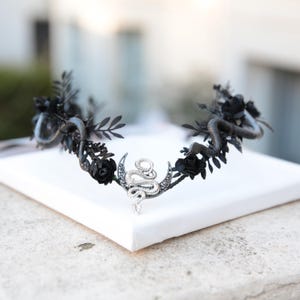May include: A gothic-style crown featuring black roses, leaves, and two snakes. The snakes are wrapped around the crown, and a silver crescent moon with a snake design is in the center. The crown is displayed on a white surface.