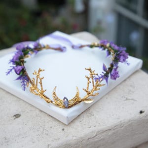 Puede incluir: Una corona dorada con un diseño de media luna y un cristal morado. La corona está adornada con flores moradas y hojas verdes. La corona está sobre una superficie blanca.