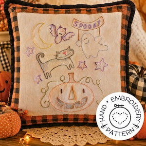 Op de afbeelding: Een vierkant sierkussen met een zwart-oranje ruitjesrand. Het midden is voorzien van handborduurwerk van een halve maan, een vleermuis, een geest met een bordje met de tekst SPOOKY, een kat, sterren en een pompoen op een crèmekleurige stof.