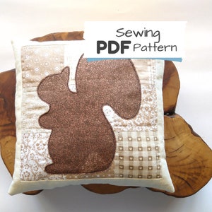 Könnte beinhalten: Ein beige-braunes Patchwork-Kissen mit einer braunen Eichhörnchen-Silhouette. Das Kissen liegt auf einer Holzoberfläche. Der Text "Sewing PDF Pattern" befindet sich auf einem blauen Banner über dem Kissen.